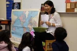 Kindergarten teacher Shizue Sommer of International Charter Academy of Georgia, a Japanese woman with light skin and dark hair, sits aside a map in front of a diverse group of young students with a large smile while holding up a paper for them to see.